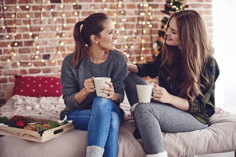lächelnde Freunde sitzen auf dem Bett und unterhalten sich bei einem heißen Getränk