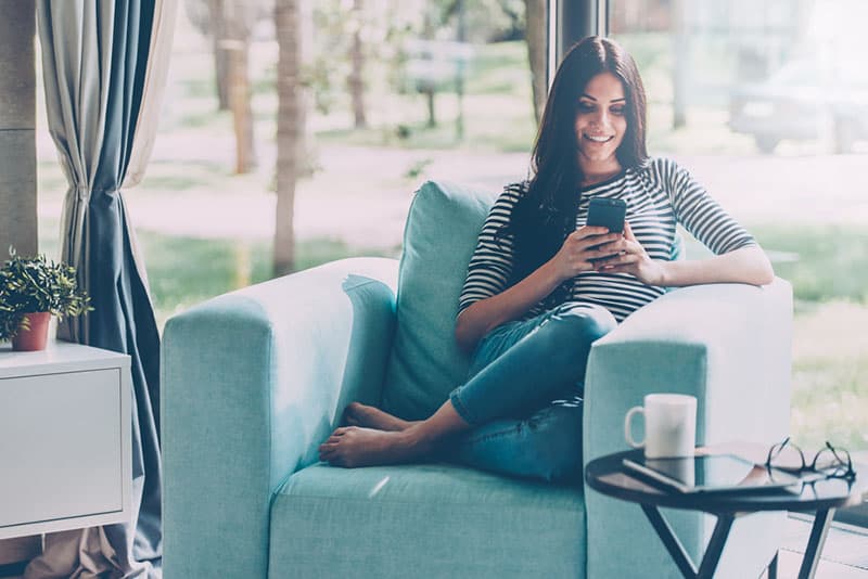 glückliches Mädchen, das auf Sofa sitzt und am Telefon tippt