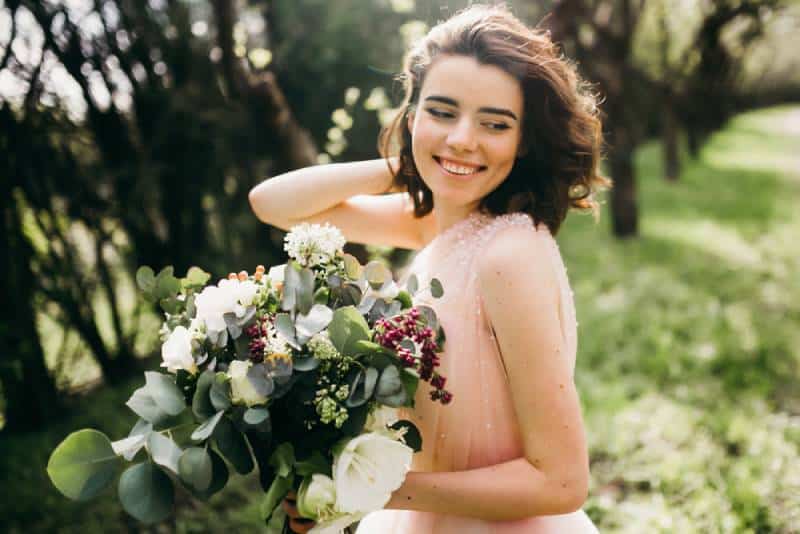 Elegante Braut, die mit Blumenstrauß im Park lächelt