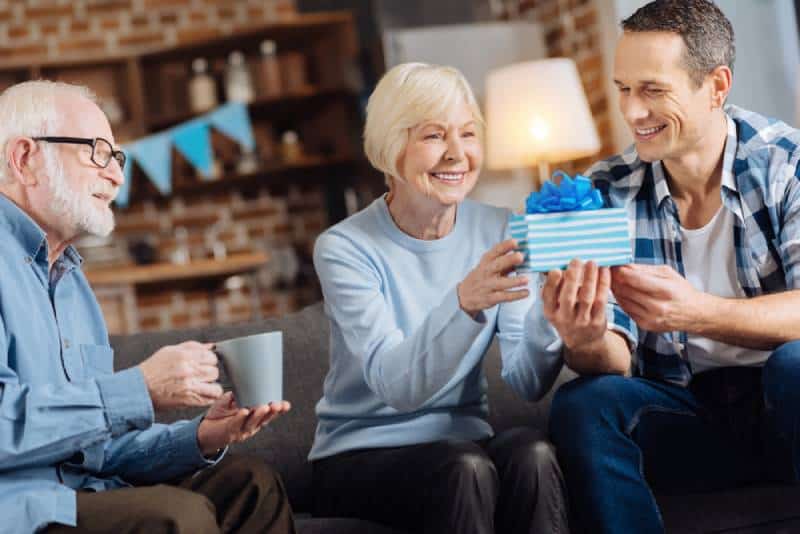 Fröhlicher junger Mann, der seiner glücklichen älteren Mutter ein Geburtstagsgeschenk gibt
