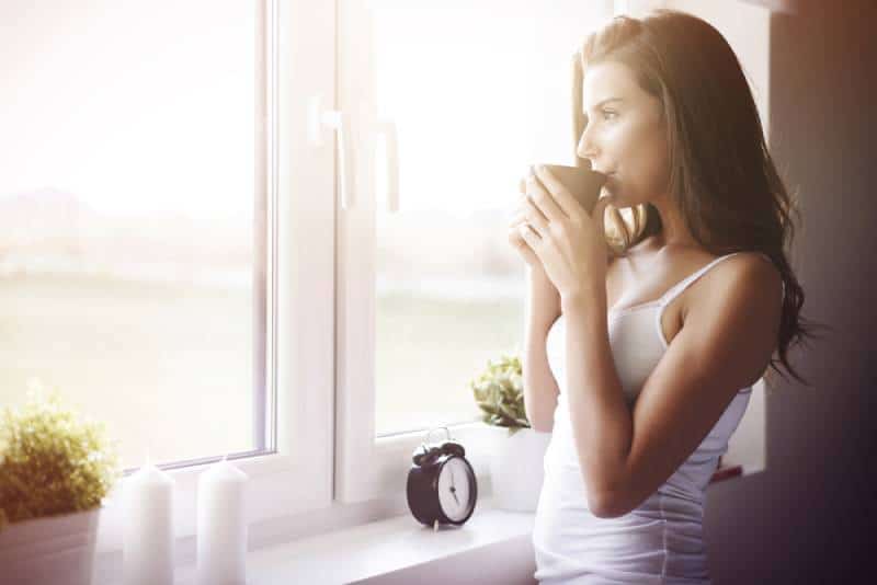 Frau, die vor dem Fenster steht und am Morgen Tee trinkt