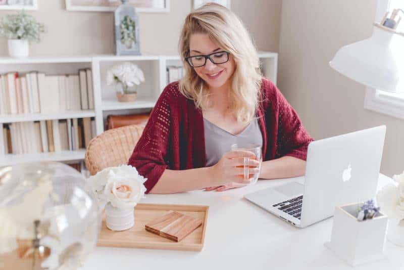 Frau lächelnd hält Glasbecher sitzend neben Tisch mit MacBook
