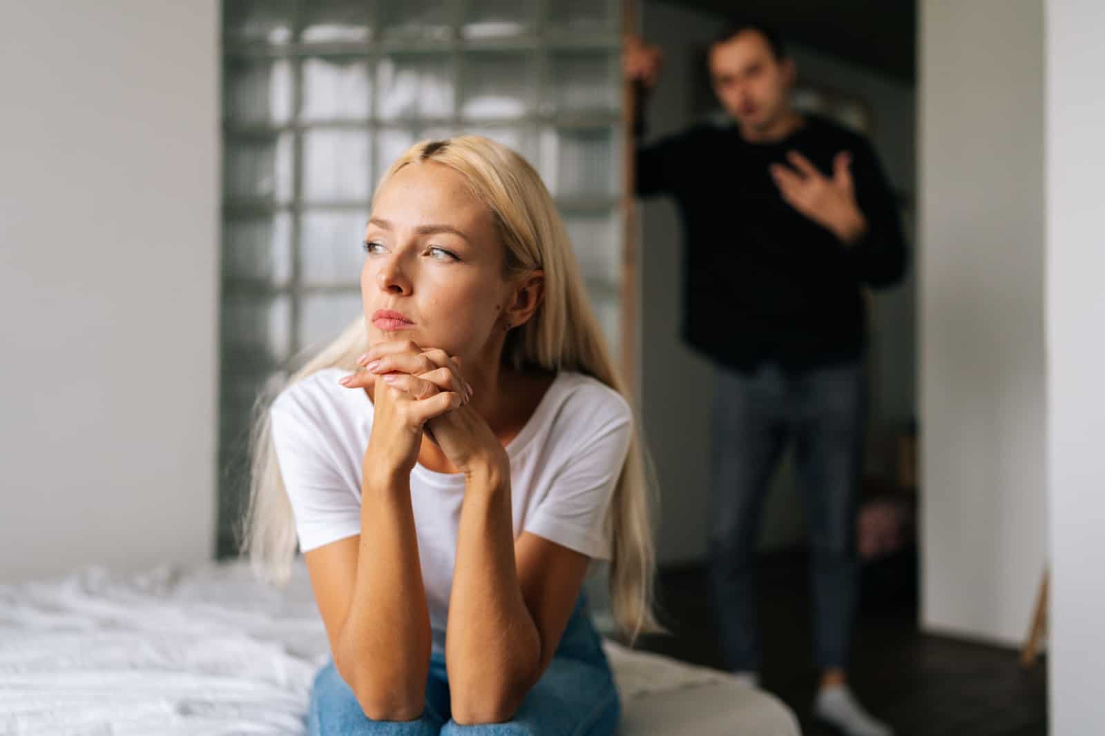 Gestresste Frau, die wegschaut, sitzt auf dem Bett auf verschwommenem Hintergrund, während der aggressive Ehemann schreit