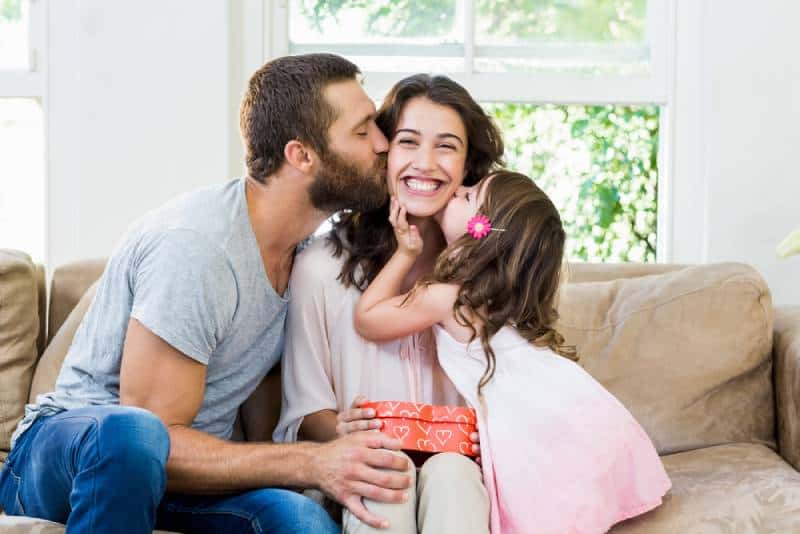 Glückliche Mutter, die ein Geschenk von ihrem Mann und ihrer Tochter im Wohnzimmer erhält