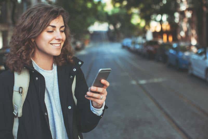 Hübsche lächelnde Frau mit Handy, das auf der Straße geht