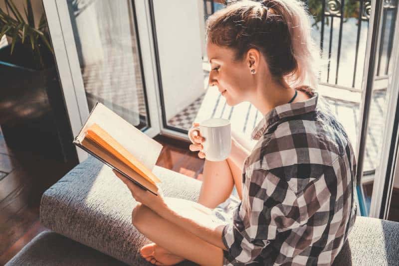 Junge hübsche Frau, die am geöffneten Fenster sitzt und Kaffee trinkt
