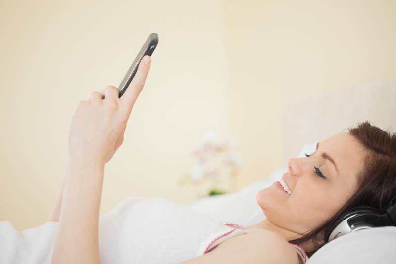 Lächelndes Mädchen, das Musik auf ihrem Smartphone hört, das auf Bett in einem Schlafzimmer liegt