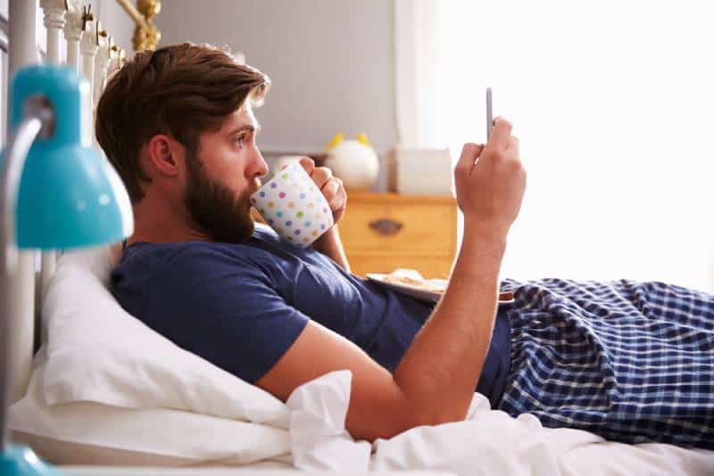 Mann im Pyjama, der im Schlafzimmer liegt und auf seinem Telefon tippt