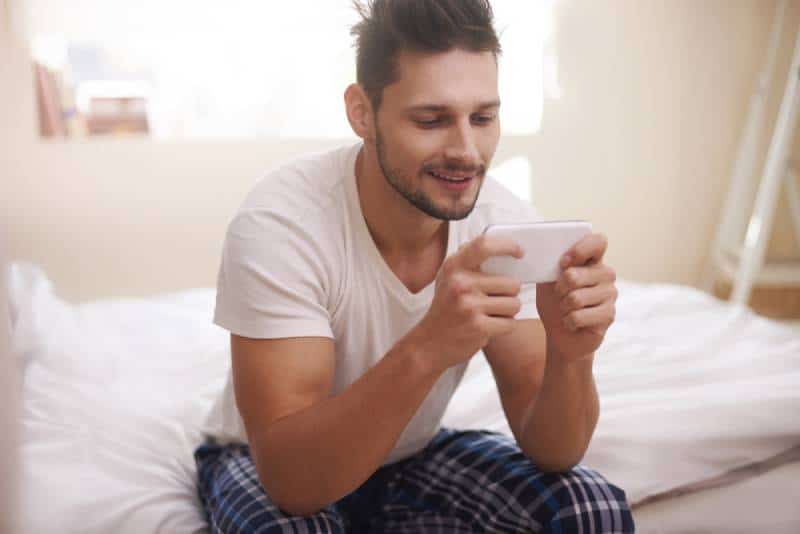 Mann sitzt auf dem Bett und tippt morgens auf seinem Handy
