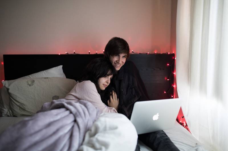Paar auf dem Bett mit MacBook Air neben dem Fenster mit weißem Vorhang