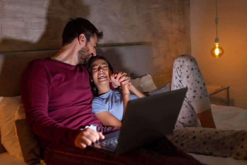 Schönes Paar, das nachts im Bett liegt, einen Laptop benutzt und sich entspannt