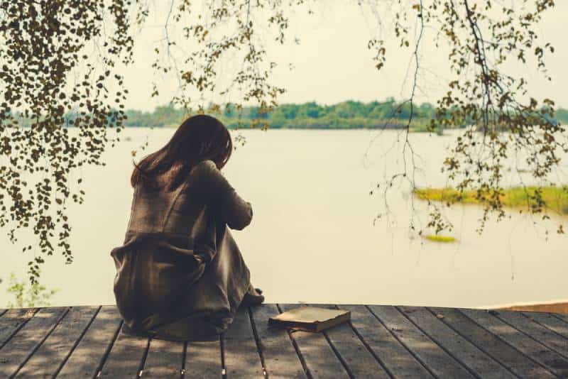 einsames Mädchen, das auf Holz am See sitzt