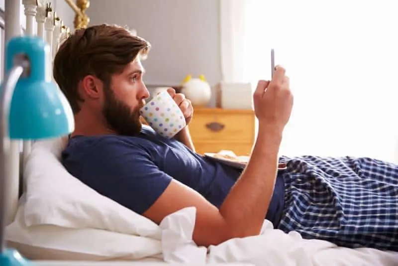 man in pajamas lying in bedroom and typing on his phone
