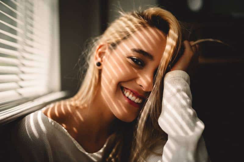 selektive Fokusfotografie der lächelnden Frau, die ihr Haar neben Jalousien hält