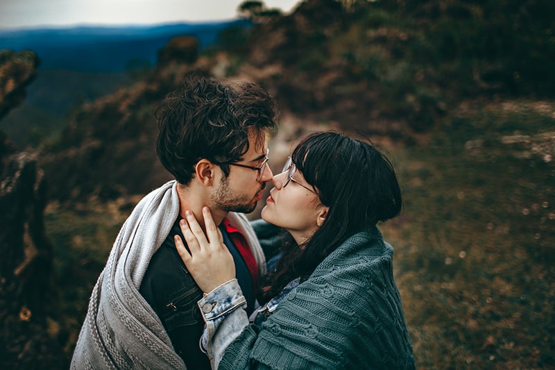 Ein Mann und eine Frau küssen sich wie Eskimos, während sie in der Natur stehen und mit einer Decke bedeckt sind