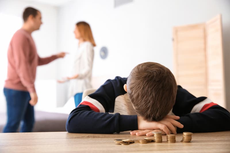 Ein trauriger kleiner Junge, der sich mit Münzen und Eltern, die sich hinter ihm streiten, auf den Tisch lehnt