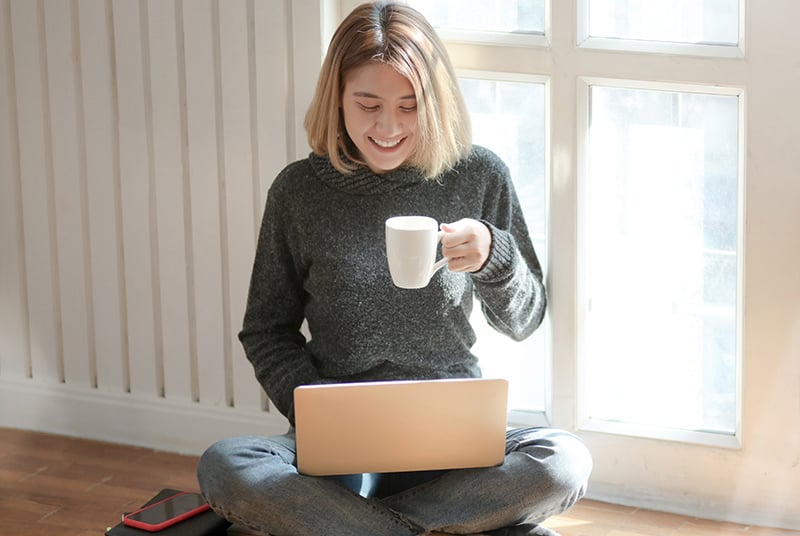 Eine Frau plaudert auf einem Laptop, während sie auf dem Boden sitzt und eine Tasse hält