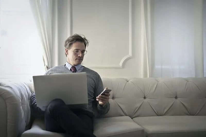 Ein Mann, der gleichzeitig Laptop und Smartphone benutzt, während er auf dem Sofa sitzt