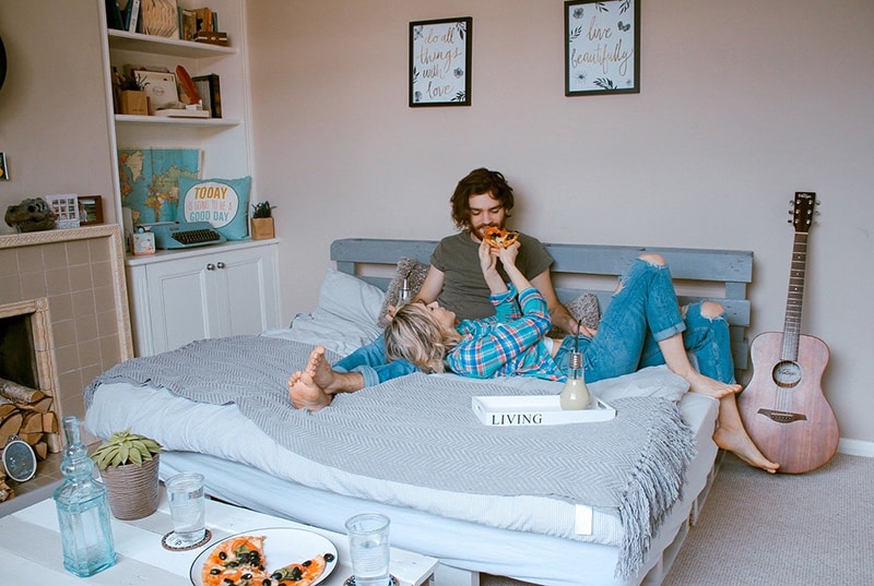 eine Frau füttert einen Mann mit Pizza, während sie auf dem Bett sitzt