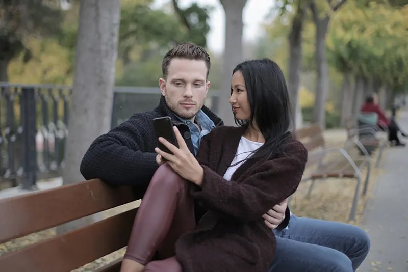 eine Frau und ein Mann sitzen zusammen auf der Bank, während eine Frau ein Smartphone benutzt
