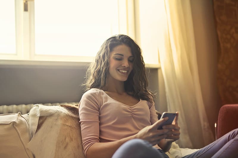 lächelnde Frau, die auf dem Sofa sitzt während der Verwendung des Smartphones