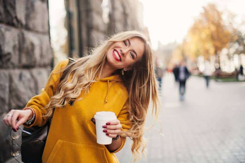lächelnde Frau im orange Sweatshirt draußen hält eine Tasse