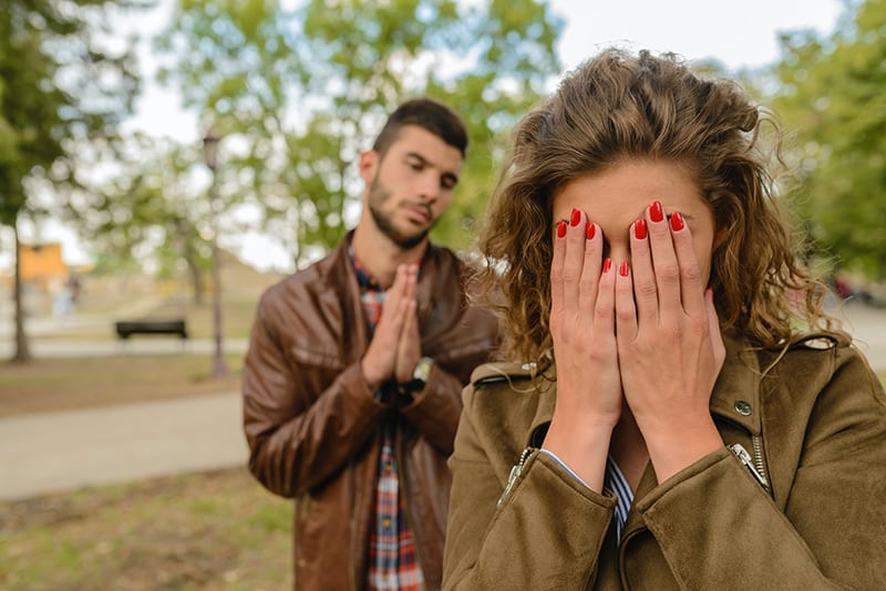 eine Frau, die ihr Gesicht mit Handflächen bedeckt, während ein Mann hinter ihr um Vergebung bittet