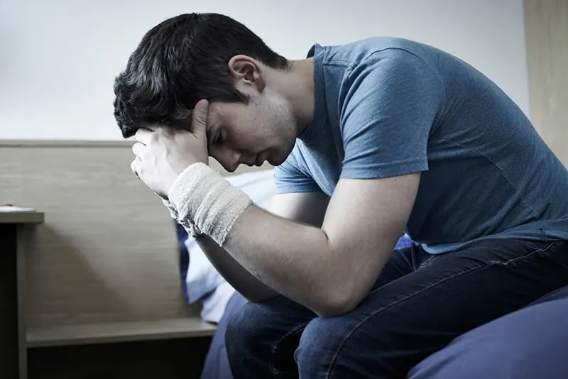 Depressiver junger Mann mit verbundenen Handgelenken nach Selbstverletzung auf der Couch sitzen