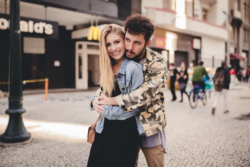 Ein Mann mit Bart hinter dem Rücken umarmt seine lächelnde Frau auf der Straße