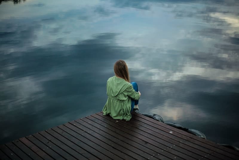 Eine Frau in einem grünen Mantel sitzt auf einem hölzernen Pier und beobachtet den See