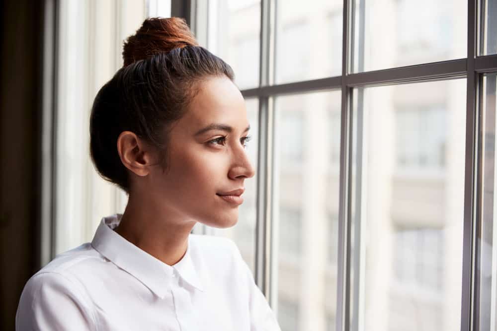 Eine Frau mit zusammengebundenen Haaren in einem weißen Hemd schaut aus dem Fenster