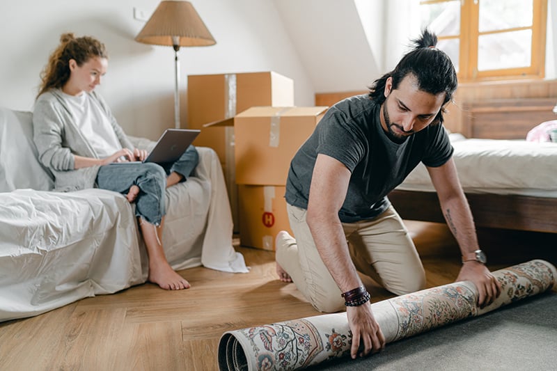 Eine Frau sitzt auf der Couch, während ein Mann einen Teppich in eine Wohnung legt