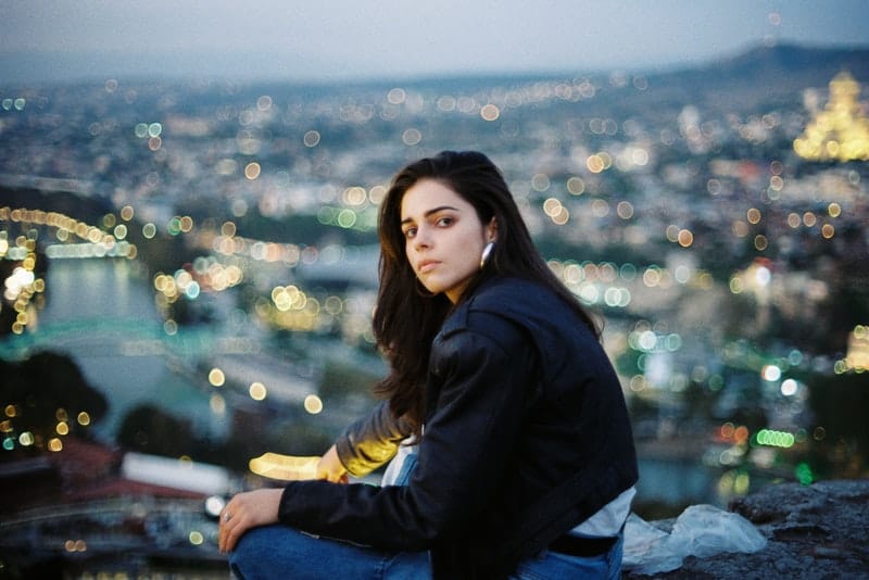 Eine ernsthafte junge Frau in einer schwarzen Jacke sitzt auf einem Felsen über der Stadt