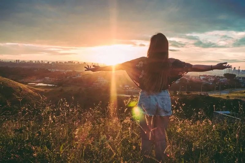 Eine Frau in kurzen Hosen steht mit ausgestreckten Armen bei Sonnenuntergang auf einem Hügel
