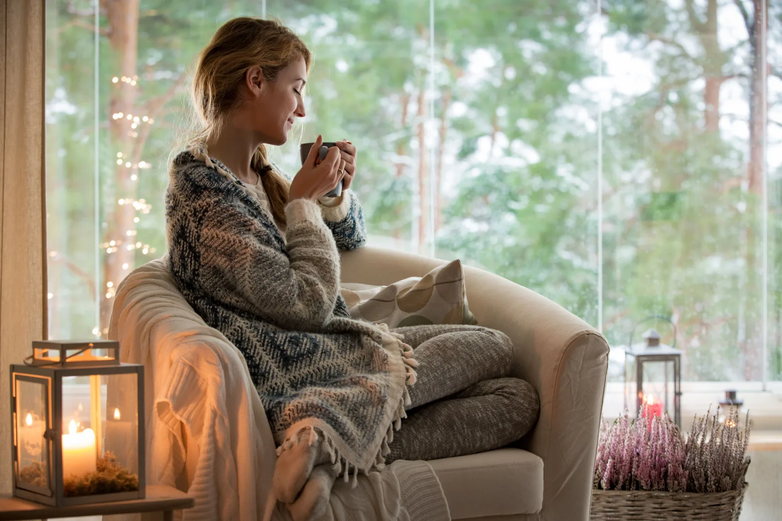 Junge schöne Frau sitzt zu Hause auf dem Stuhl am Fenster mit einer Tasse heißen Kaffee