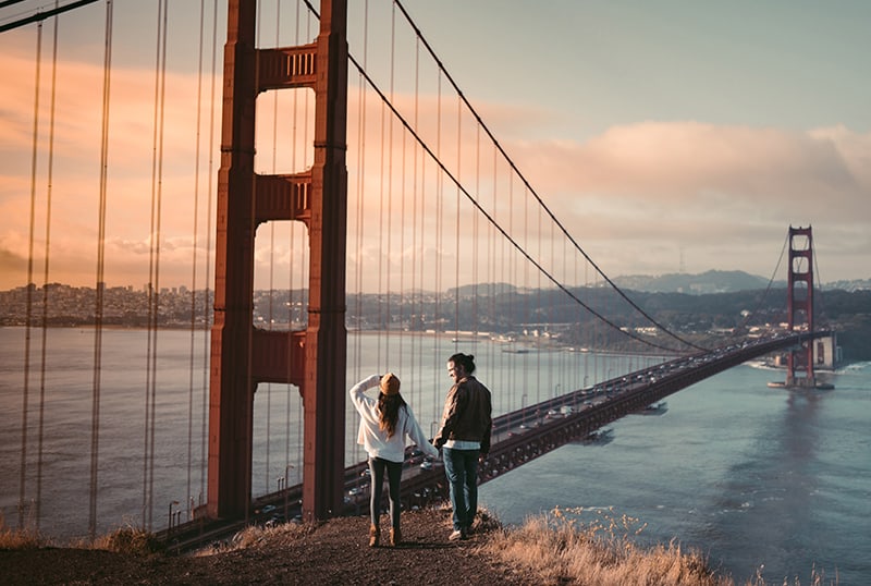 ein Paar Händchen haltend, während es in der Nähe der Golden Gate Bridge steht und den Sonnenuntergang beobachtet