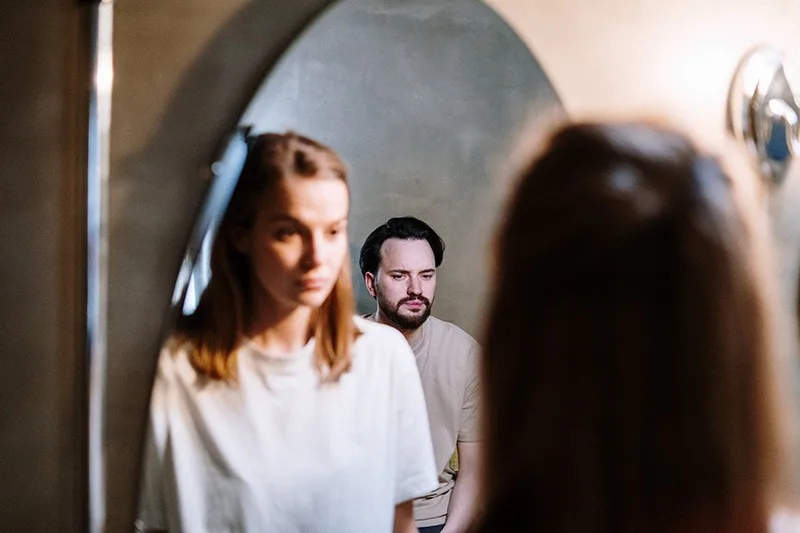 Ein emotional verletzter Mann sitzt hinter einer Frau, während sie vor dem Spiegel steht
