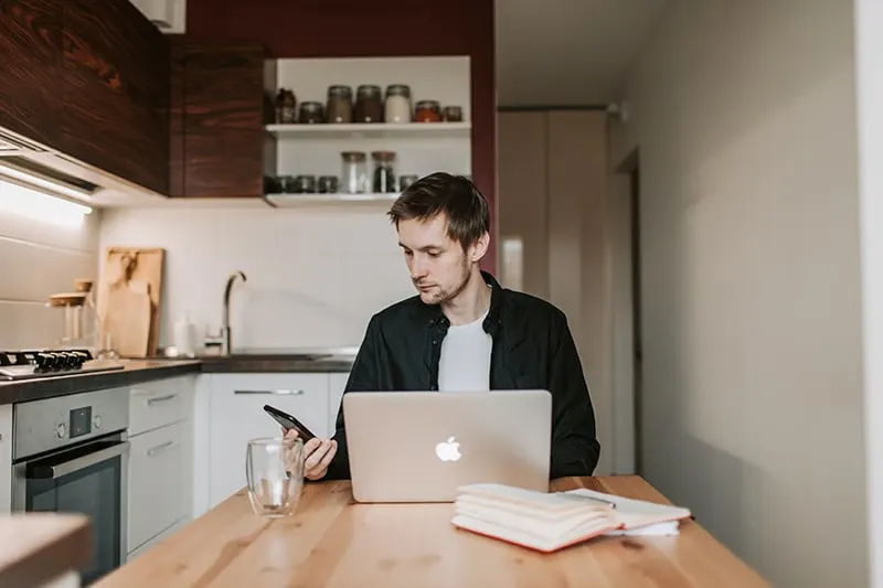 ein nachdenklicher Mann, der ein Smartphone betrachtet, während er vor Laptop sitzt