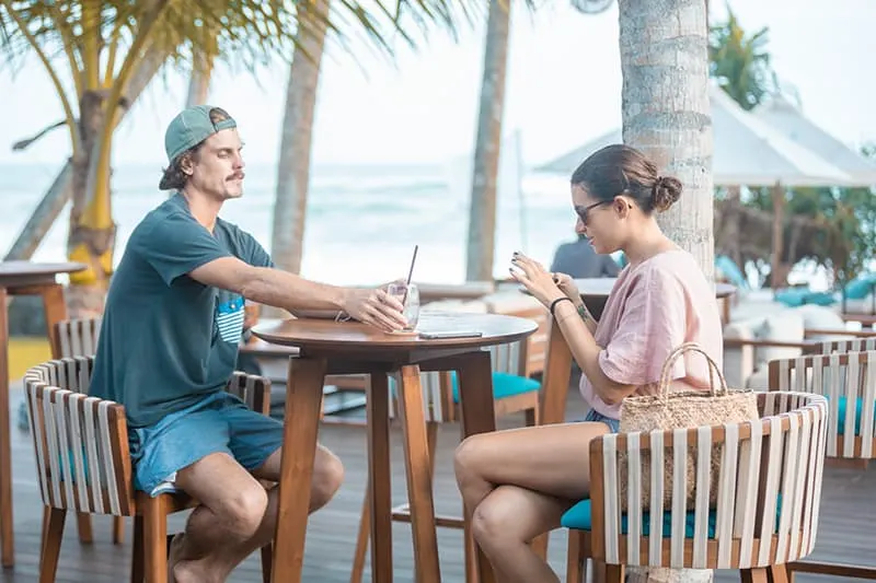 eine Frau, die ein Smartphone benutzt, und ein Mann, der ein Glas hält, während sie in einem Café draußen sitzen