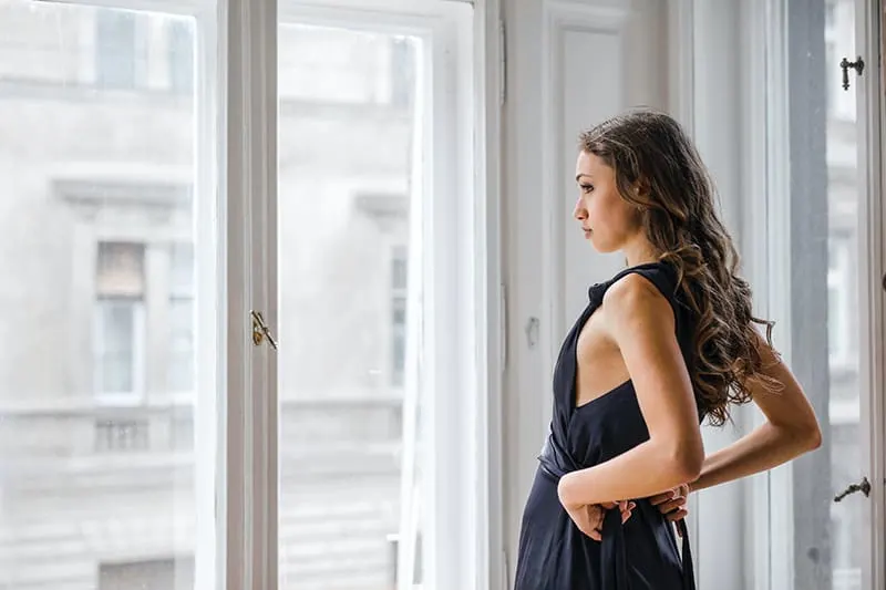 eine nachdenkliche Frau, die neben einem Glasfenster steht, während sie ein Kleid anzieht