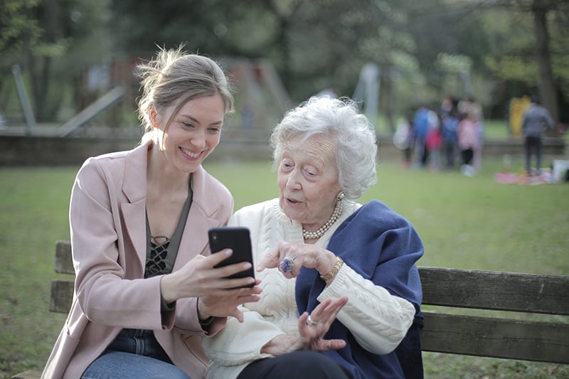 fröhliche Großmutter und erwachsenes Enkelkind, die ein Smartphone während eines Gesprächs beim Sitzen auf der Bank betrachten