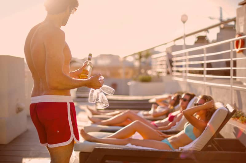 junger Mann bietet sonnenbadende freundinnen am schwimmbad getränke an