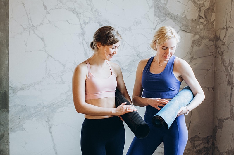 Zwei Frauen vergleichen ihre Yogamatten, während sie in der Nähe einer grauen Wand stehen