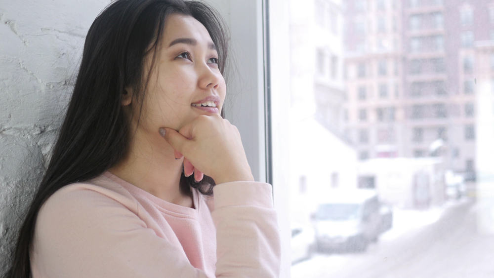 Die asiatische Frau mit einem Lächeln denkt nach und sitzt am Fenster