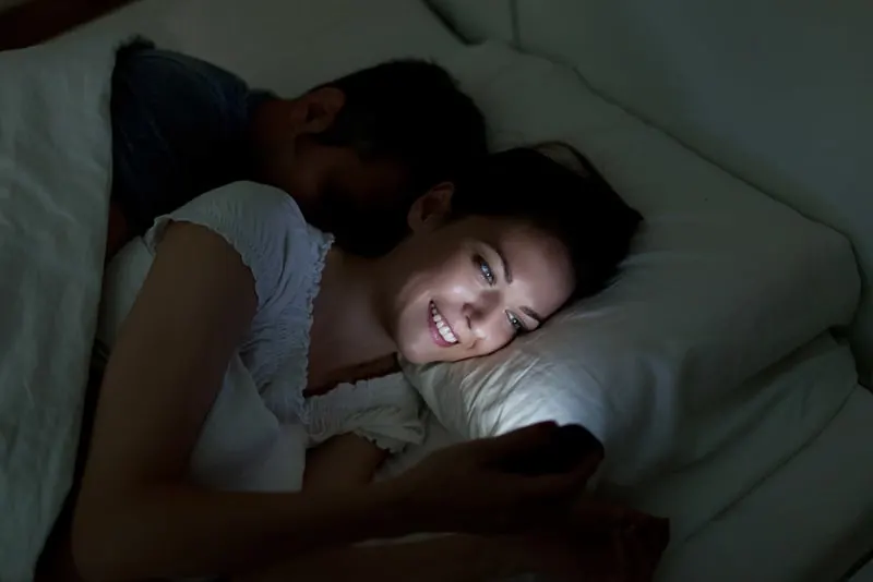 eine lächelnde Frau, die ein Smartphone in einem Bett benutzt, während ihr Mann hinter ihr schläft