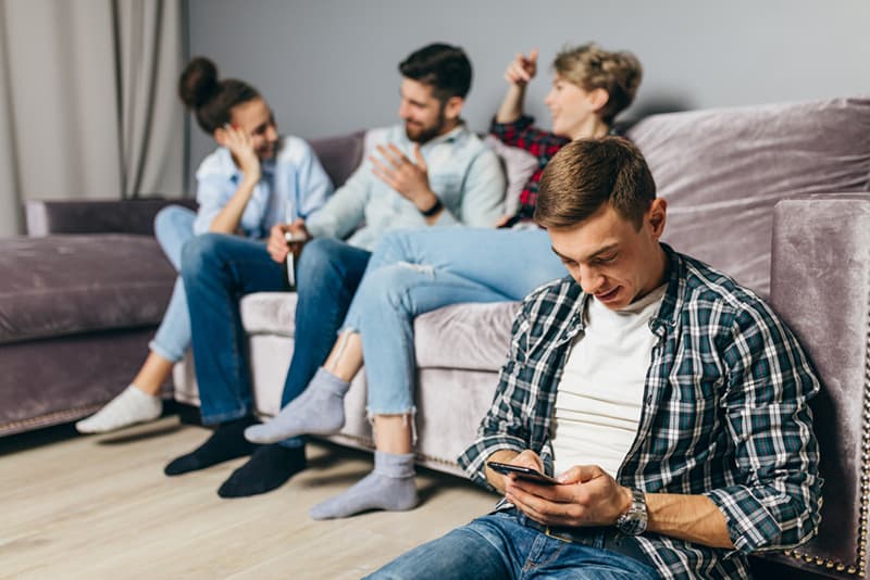 Ein Mann sitzt auf dem Boden und benutzt ein Smartphone, während ihre Freunde reden und auf der Couch sitzen