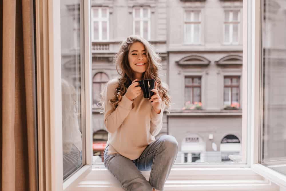 Ein glückliches Mädchen mit schönen langen Haaren sitzt am Fenster und trinkt Kaffee
