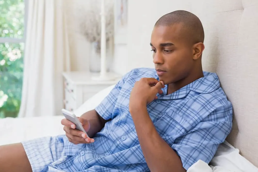 Ein schwarzer Mann im blauen Pyjama liegt auf dem Bett und benutzt ein Smartphone