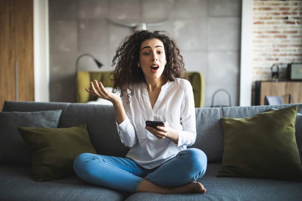 Eine überraschte Frau in einem weißen Hemd mit einem Handy in der Hand sitzt auf der Couch