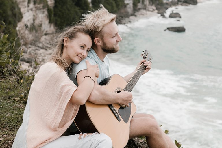 Eine Frau umarmt ihren Mann von hinten, während er auf den Felsen in der Nähe des Meeres Gitarre spielt